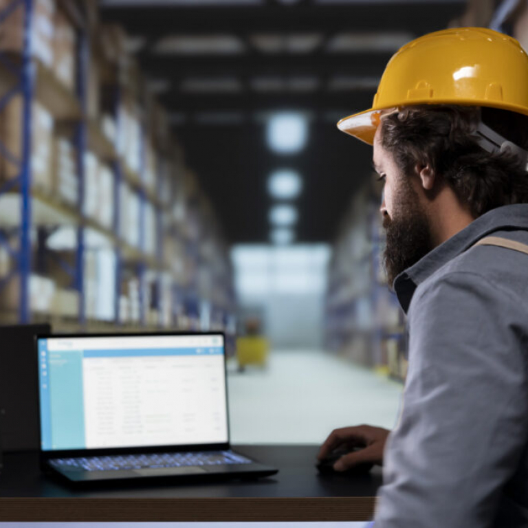 Worker using a laptop in a distribution warehouse, illustrating how a Sage 200 warehouse management system supports inventory tracking and operational efficiency.