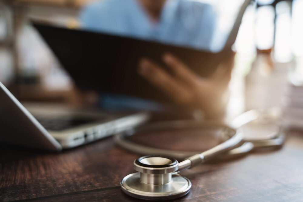 Close-up of a stethoscope on a desk with a healthcare professional reviewing financial reports on a tablet, illustrating the use of financial management software in healthcare.